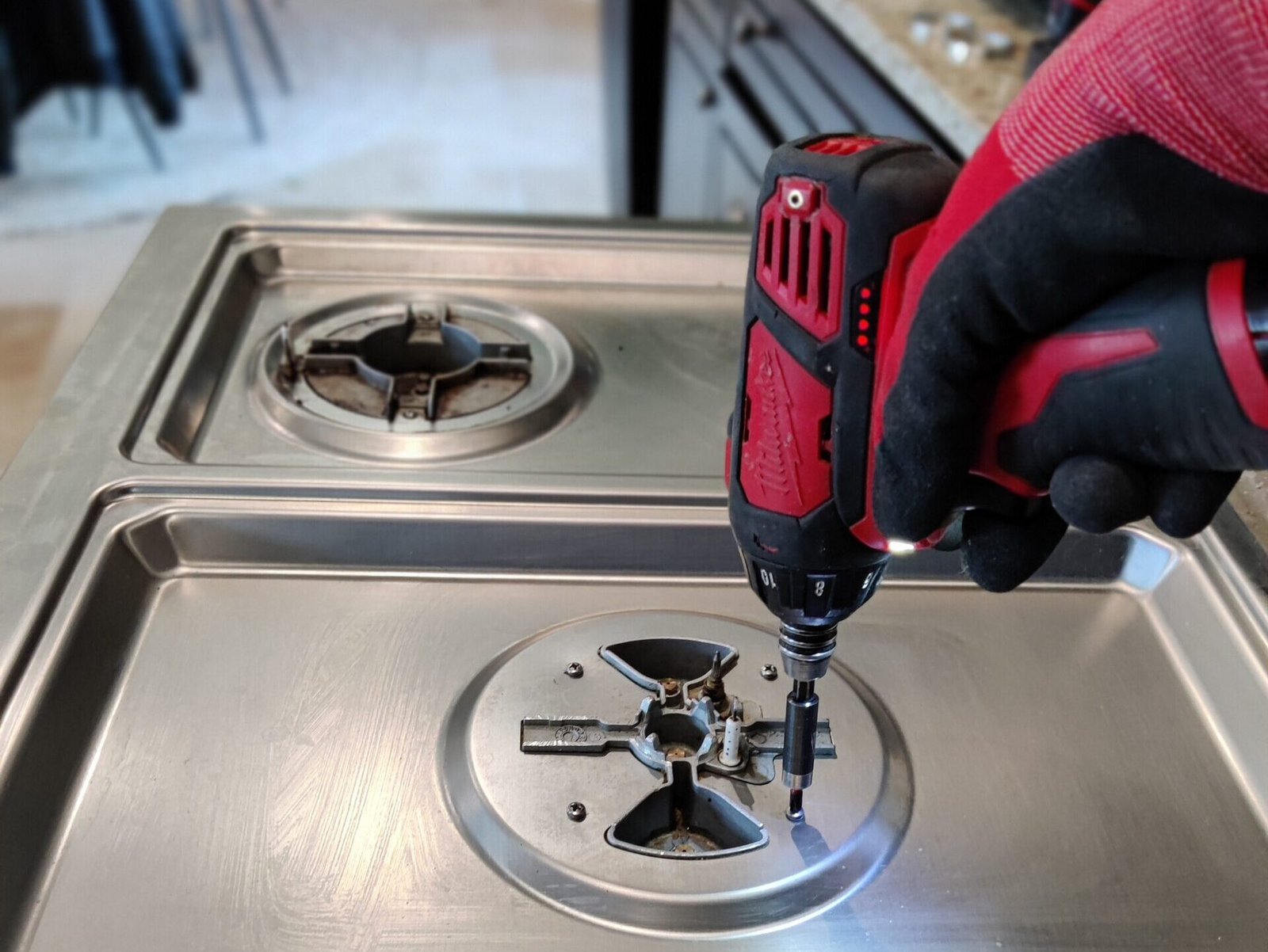 Appliance technician using a power tool to dismantle and repair a sealed gas burner on a Wolf stainless steel cooktop.